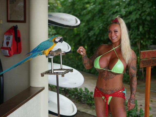 BonnievonClyde-hot with big boobs, green bikini, tattoos, blonde ponytail, red headband, holding food, standing next to blue and yellow macaw on a scale.