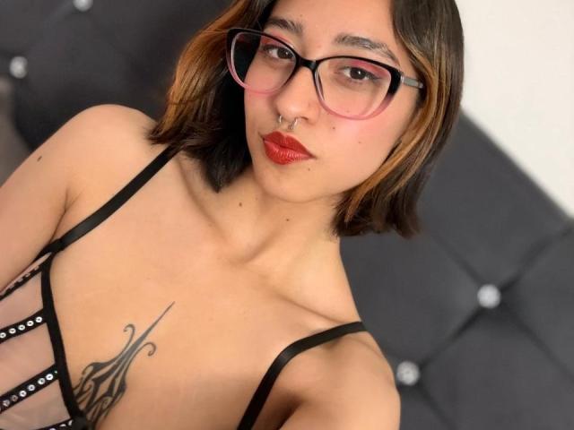 CamiVargas with glasses, red lipstick, and a nose ring, wearing a black strappy top, showing a tattoo under her tits. She has a medium skin tone and a bob.