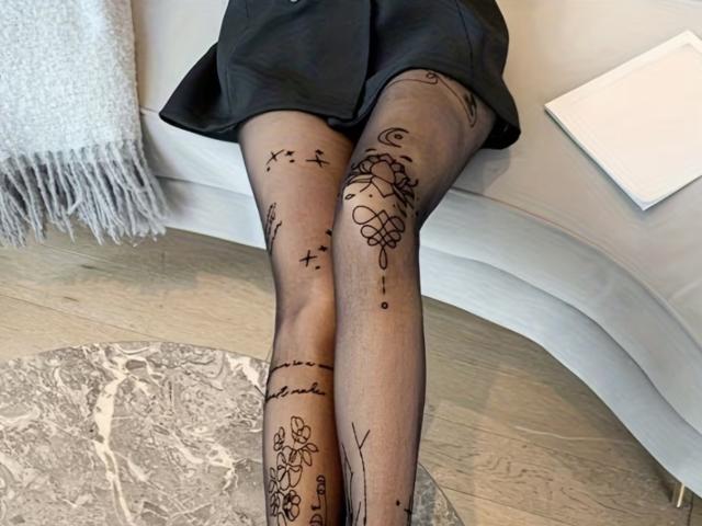 MargoFun's legs in sheer black tights with black line tattoos, sitting on a white curved surface, wearing a black skirt, with a white textured rug underneath.