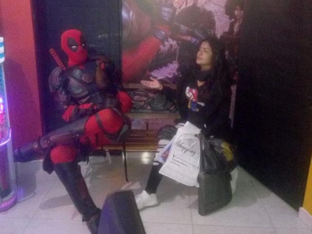 ArianithaXox in red and black Deadpool costume sits on a chair with leg raised, looking at a girl with curly hair, black shirt, and white bag. She's on tiled floor.