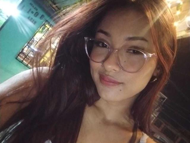 SashaHans with long brown hair, wearing clear glasses, smiling at the camera, showing her small nose and light pink lips. Her tits are slightly visible under a tank top.