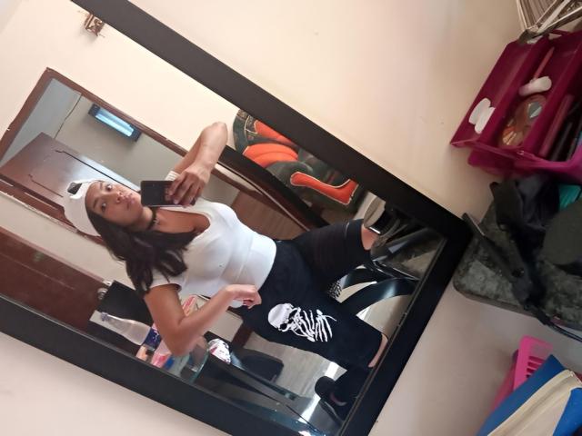 ArianithaXox in a white crop top, black skull-patterned pants, and black boots, standing in front of a mirror, with a white cap, showing her tits and ass.