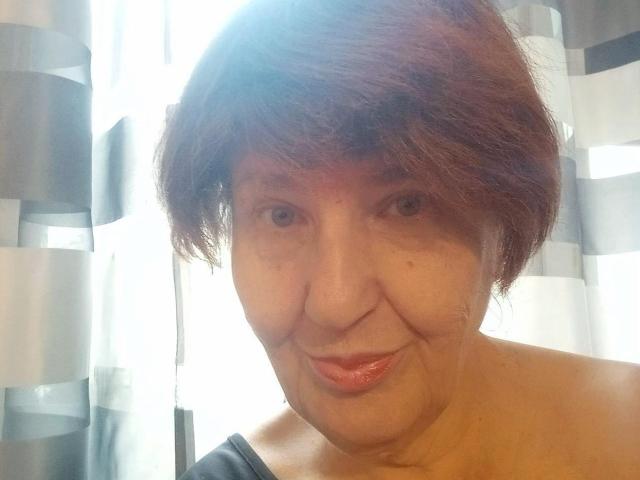 MioritaStar has short brown hair, a wrinkled face, and a small smile with red lipstick, wearing a black strap, showing her ass and a bit of her tits.