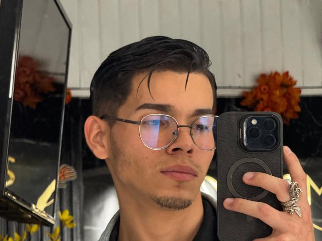 SmileKai with short black hair, glasses, and a ring, taking a selfie with a black phone in a mirror. Reflective eyes, subtle beard, and a confident smile.