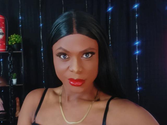 VenusMiller with long black hair, red lipstick, and blue eyes, wearing a black top and gold necklace, looking at the camera with a slight smile. Her tits are small.