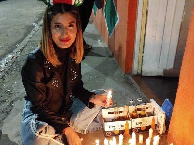 Kaiix with shoulder-length blonde hair, black leather jacket, and blue jeans, kneeling, holding a candle, in front of a box of Corona beer candles on a sidewalk.