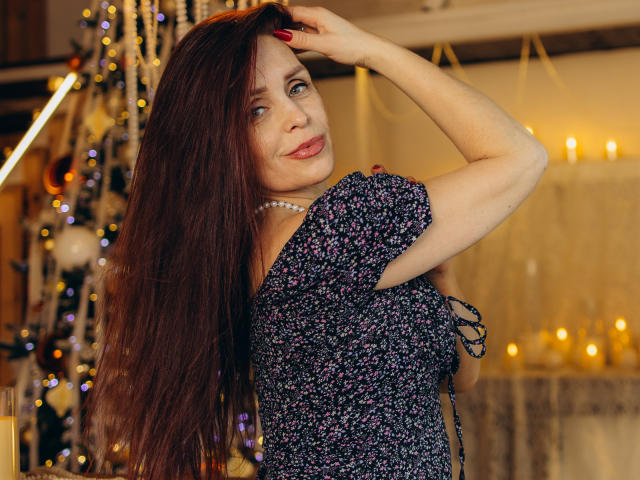 AlexisLovelyy with long brown hair, wearing a floral dress, pearl necklace, and red nails, touches her head, smiling, candles in the background, soft warm lighting, attractive.