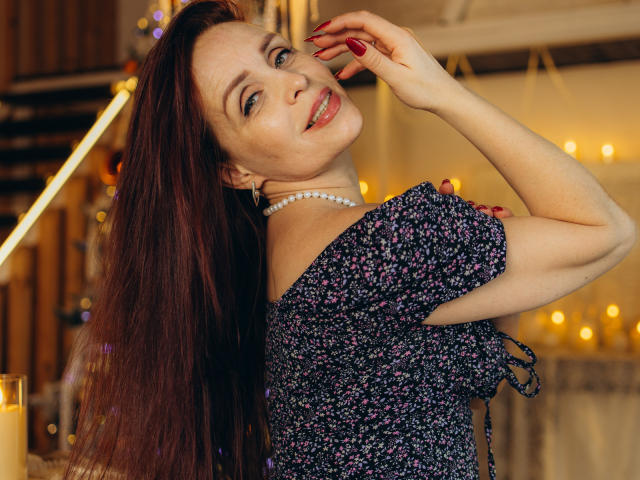 AlexisLovelyy with long red hair, smiling, wearing a floral dress, pearl necklace, red nails, raising her right hand, showing off her tits, in a warmly lit room.