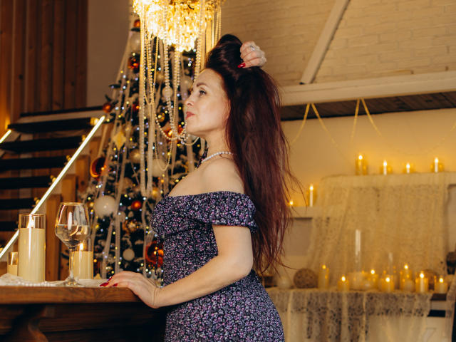AlexisLovelyy with long brown hair, wearing an off-shoulder floral dress, touching her hair, standing by a table with a glass of wine, surrounded by candles and a Christmas tree.