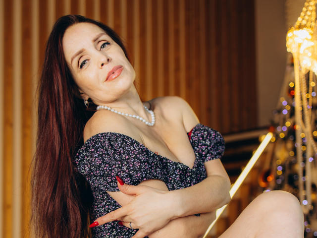 AlexisLovelyy with long red hair, wearing a floral off-shoulder dress, pearl necklace, and red nails, sitting with crossed arms, showing her tits, and a slight, sexy smile.