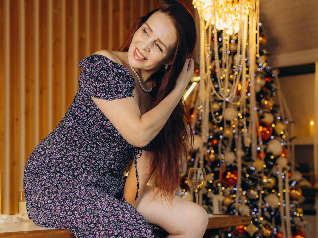 AlexisLovelyy with long red hair, wearing a floral dress, sitting on a table, smiling, one eye closed, hand in hair, pearl necklace, Christmas tree with lights in background.
