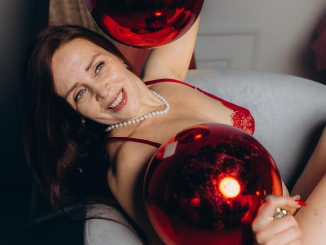 AlexisLovelyy with red bra, pearl necklace, holding shiny red orbs, smiling, showing tits, brown hair, blue eyes, light skin, sitting in gray chair, arm raised, ring visible.
