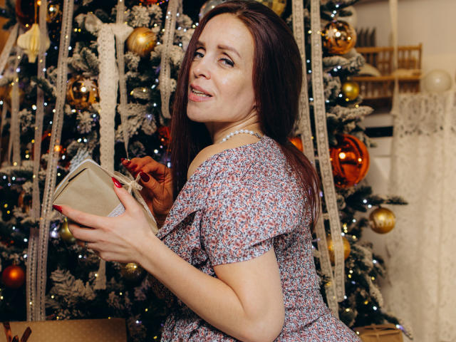AlexisLovelyy with medium tits, wearing a floral dress, holding a gift, standing in front of a Christmas tree, smiling, wearing a pearl necklace, brown hair, red nails.