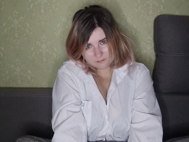 MinistraX with medium brown, slightly wavy hair, wearing a white button-up shirt, sitting on a dark gray couch, looking directly at the camera with a slightly curious expression.