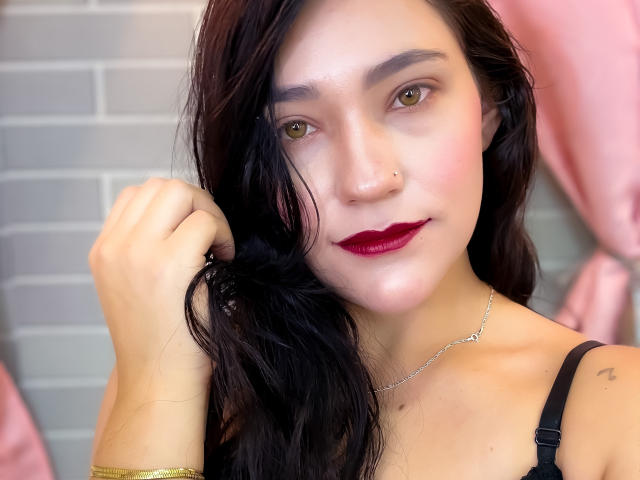 TamaraCians has fair skin, dark wavy hair, green eyes, red lipstick, black bra, silver necklace, gold bracelet, nose ring, and a small tattoo on her right shoulder.