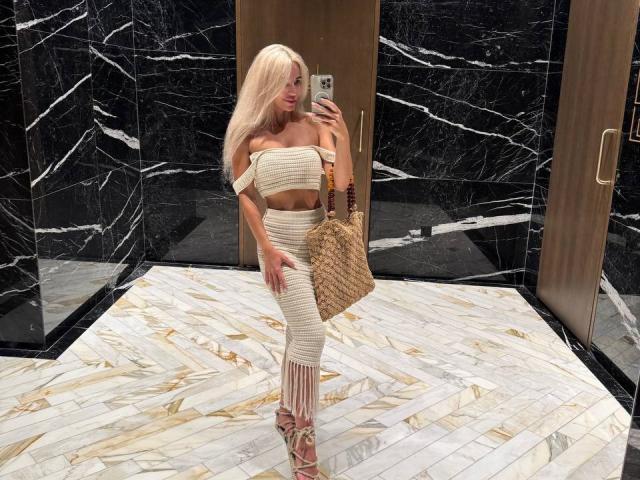 BellaRayyy with blonde hair, off-shoulder beige crop top, matching ribbed pants, holding wicker bag, taking selfie in marble-laden room, wearing strappy beige heels.