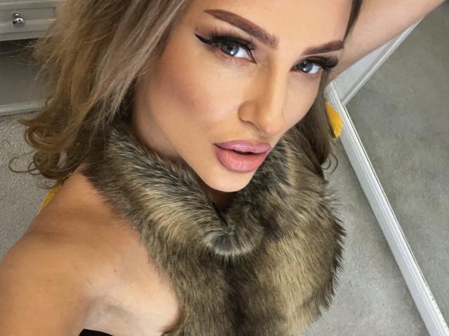 YaseminSaf with pouty pink lips, thick black eyeliner, and a furry collar, looking up with sultry eyes, blonde hair, and a tan complexion. Boobs partially visible.