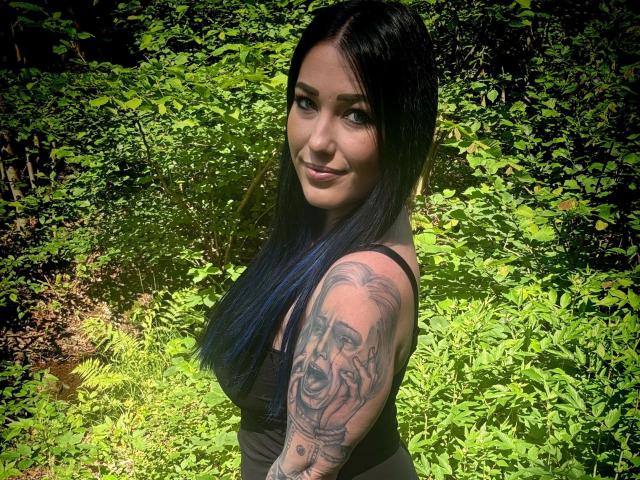 Only-Sophia-hot with long blue-black hair, black tank top, tattooed arm, smiling at camera, standing in sunlit green forest, looking sexy, cute, and confident.