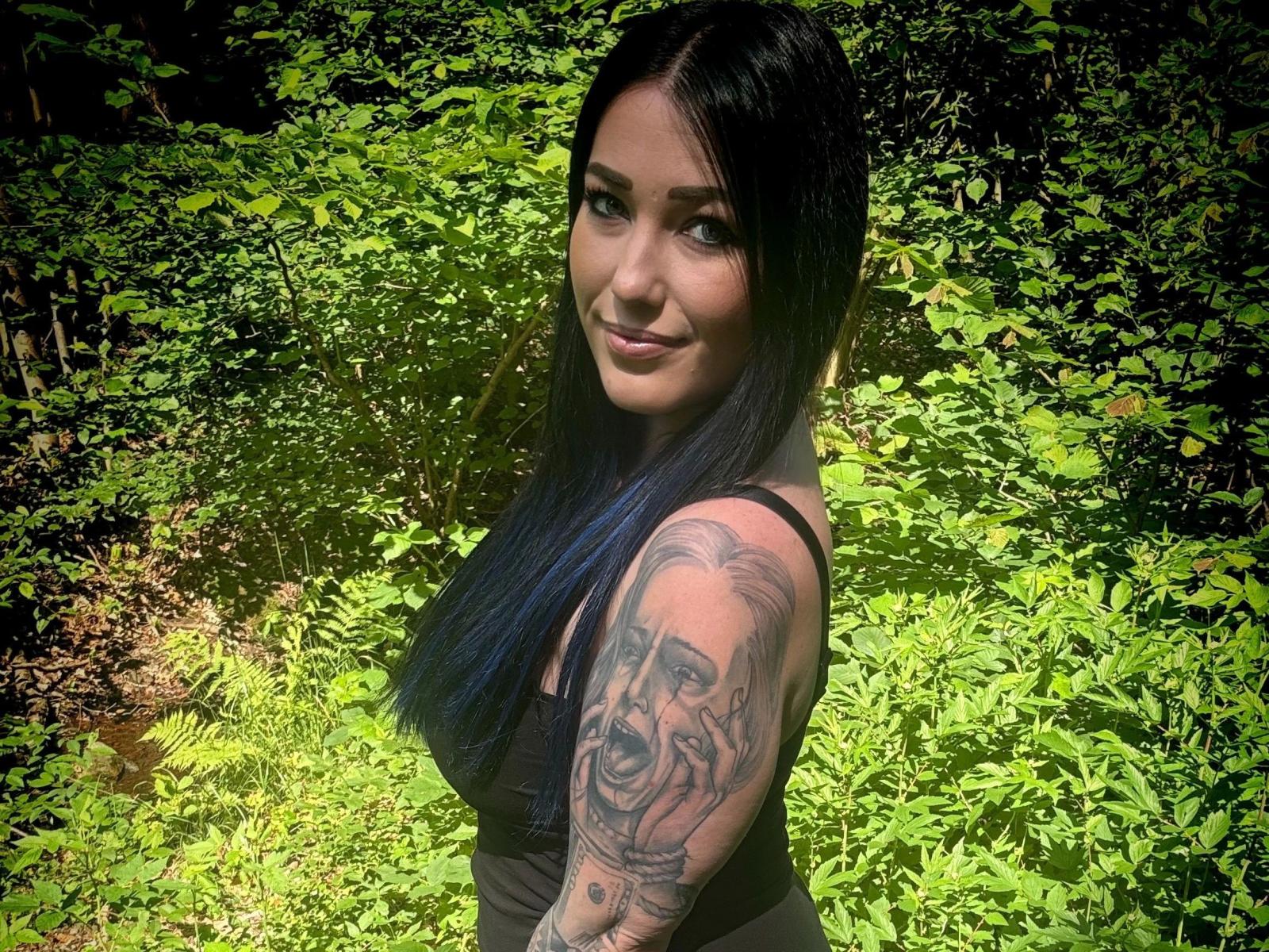 Only-Sophia-hot with long blue-black hair, black tank top, tattooed arm, smiling at camera, standing in sunlit green forest, looking sexy, cute, and confident.