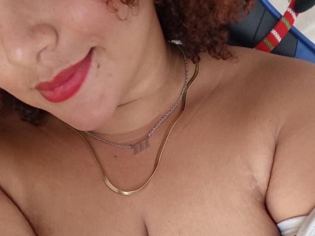 YeraCarey with curly brown hair, red lipstick, and a gold necklace, showing her bare chest and small, round tits. She's smiling slightly with a cute nose freckle.