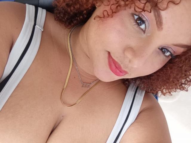YeraCarey with curly red hair, light skin, pink lipstick, blue eyeshadow, gold necklace, white tank top, showing her big boobs, smiling at the camera.
