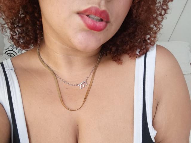 YeraCarey's face, showing red lips, curly brown hair, and a gold chain with a small "T" pendant, wearing a low-cut white top, showing her big, round tits.