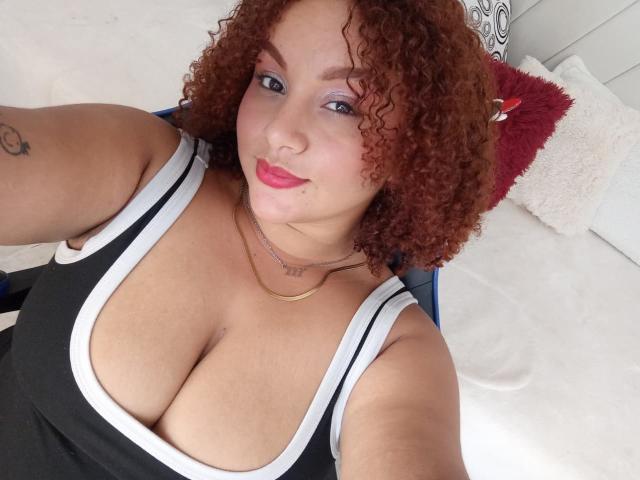 YeraCarey with curly red hair, wearing a black tank top, showing large boobs, pink lipstick, and a gold necklace, smiling at the camera.
