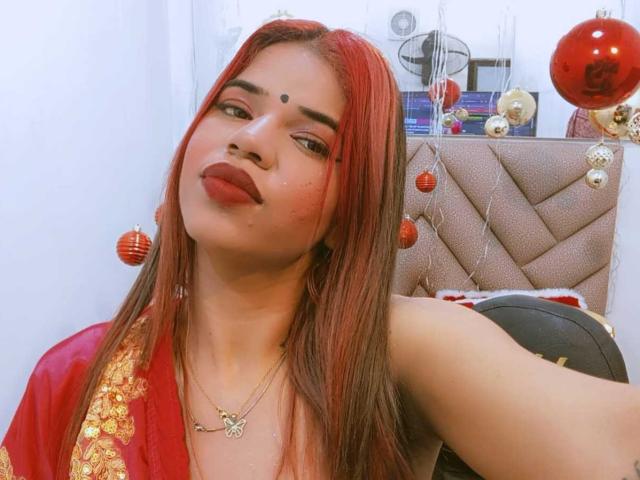 KairaDayamai with red hair, dark eyebrows, and a red bindi, wearing a red and gold top, has a cute face and a small gold necklace. She's taking a selfie.