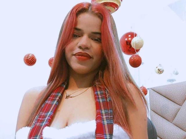 KairaDayamai with red hair, wearing a plaid scarf, white fur top, and a candy cane hat, has a cute smile and small tits, surrounded by floating Christmas ornaments.
