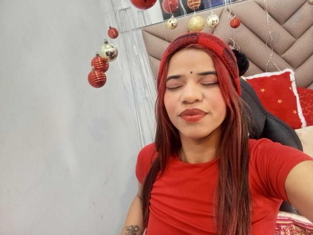 KairaDayamai with red hair, red beanie, and red shirt, smiling with eyes closed, nose piercing, and a small mole above her lip, sitting with a tattoo on her arm.
