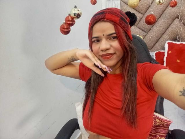KairaDayamai with red shirt, red and black beanie, red lipstick, long brown hair, tattoos on arms, hand on cheek, smiling, Christmas ornaments in background, sitting on black chair.