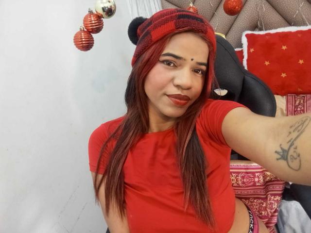 KairaDayamai with red shirt, red and black beanie, red lipstick, and red eyebrows, showing a tattoo on her right arm, in a selfie, with Christmas ornaments in the background.