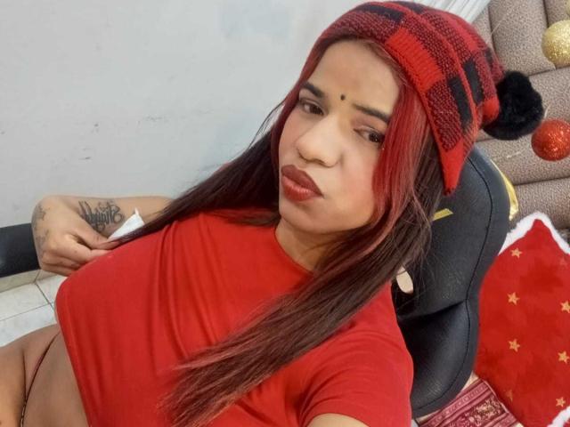 KairaDayamai with red plaid beanie, red shirt, pouty lips, tattoo on left arm, holding paper, sitting, showing her boobs, long red and black hair, red lipstick.