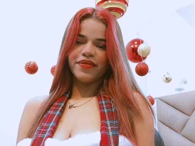 KairaDayamai has red hair, wearing a plaid vest, white dress, and red lipstick, with Christmas ornaments floating around, and a candy cane hat on her head.