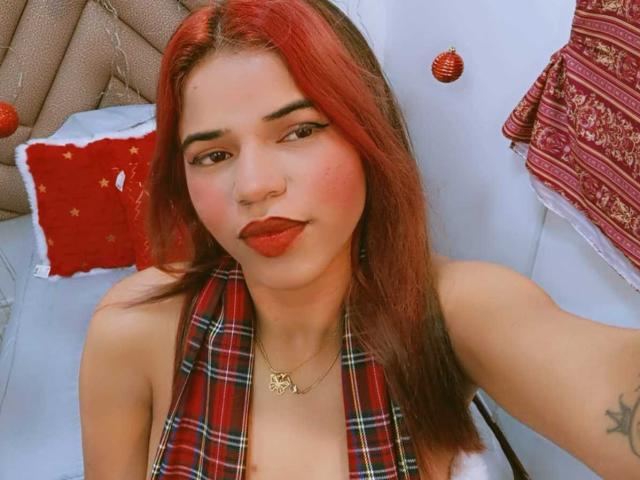 KairaDayamai with red hair, plaid scarf, red lipstick, nose ring, and necklace, smiling at the camera, showing her tits and a tattoo on her right arm.