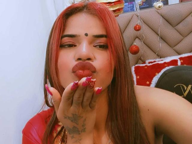 KairaDayamai with red hair, makeup, and a dot on her forehead, pouting with her hand near her red lips, showing off her pink nails and tattooed hand.
