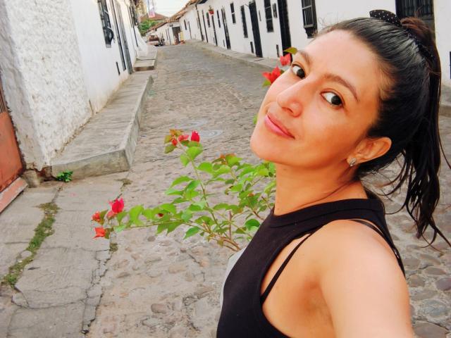 SarahFriendly with brown eyes and black hair in a ponytail, wearing a black sleeveless top, smiling at a cobblestone street with red roses in the foreground.