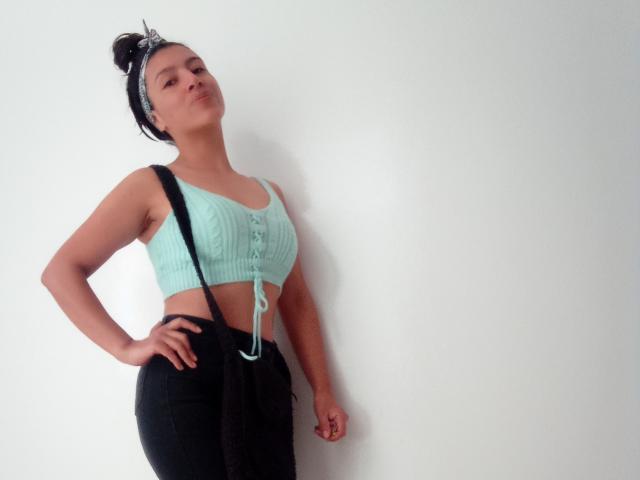 SarahFriendly a la peau mate, porte des bretelles noires, un débardeur vert menthe à rayures avec des boutons, et un pantalon noir. Ses cheveux sont attachés en chignon avec un bandeau noir..