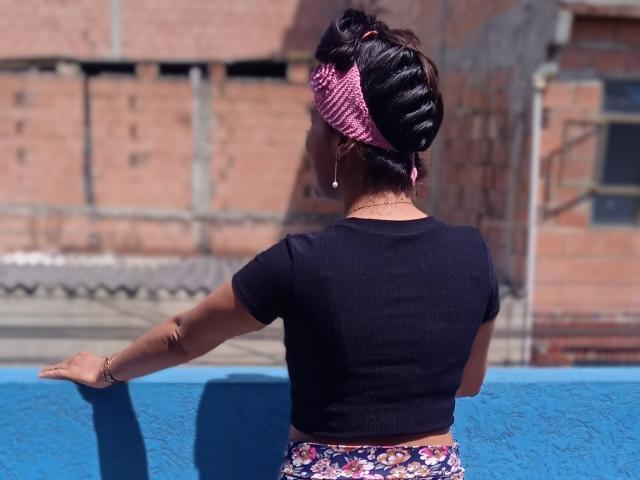 SarahFriendly è di nuovo qui, indossa una maglietta nera e una gonna floreale, un foulard rosa, ha le braccia aperte, sullo sfondo c'è un muro blu e un edificio in mattoni, c'è luce solare, i suoi capelli scuri sono raccolti in uno chignon..