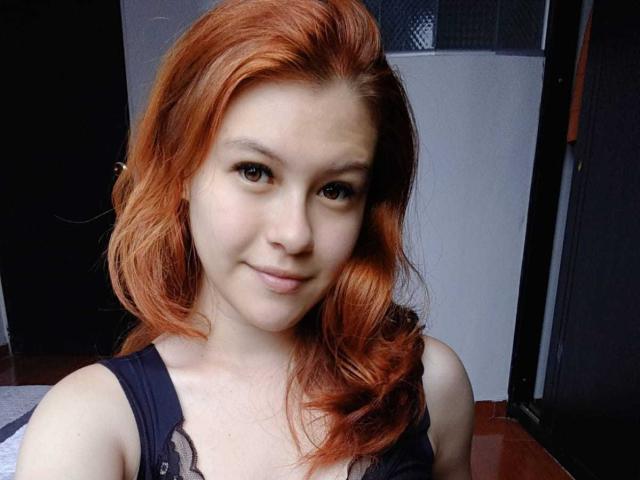 TaylorCarpert with red hair, small tits, fair skin, and a slight smile, wearing a black tank top, in a casual selfie.