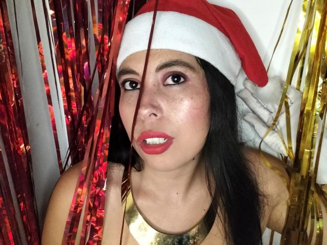 NikkieMilky with black hair, red lipstick, and a Santa hat, wearing a gold necklace, standing between red and gold streamers, looking at the camera with a slightly open mouth.