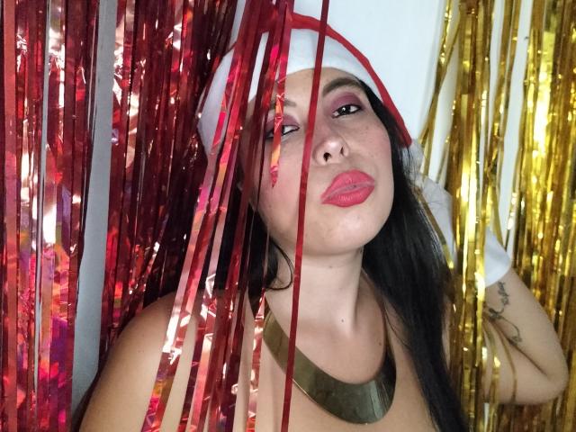 NikkieMilky with red and gold metallic streamers in front of her face, wearing a red and white hat, gold necklace, and red lipstick, showing tattoos on her right arm.