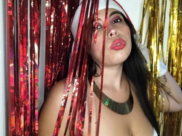 NikkieMilky with red and gold metallic strips in front, wearing a white hat and metallic necklace, has pink lipstick and black hair, showing her tits and tattooed arm.