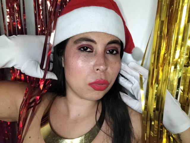 NikkieMilky with red Santa hat, white gloves, and red lipstick, has brown eyes, freckles, and glossy pink lips, wearing a gold top, with shiny red and gold streamers.