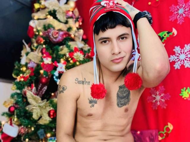 Shirtless KeyMcKinley with red and white hat, puffy red pom-poms, tattoos, black wristwatch, short black hair, brown eyes, slight smile, Christmas tree, red background, and ornaments.