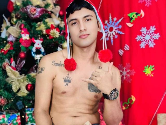 Shirtless KeyMcKinley with a Santa hat, holding a red pom-pom, has tattoos on his chest and arm, wearing a watch, standing in front of a Christmas tree.