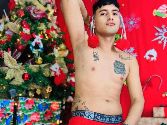 KeyMcKinley, shirtless, with a red pom-pom in his armpit, has tattoos, short black hair, and a slight smile, wearing red Calvin Klein underwear, in front of a Christmas tree.