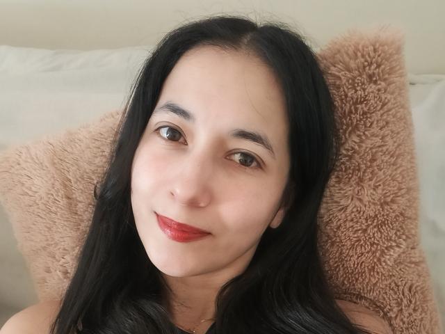 AnneStone has long black hair, brown eyes, and red lipstick, lying against a fluffy pink pillow, smiling softly. She's fair-skinned with a cute face.