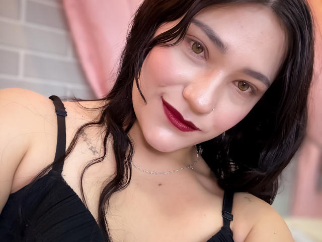 TamaraCians has light skin, brown eyes, black wavy hair, dark red lipstick, black bra, small tattoo on left shoulder, nose ring, and a silver necklace, smiling at the camera.