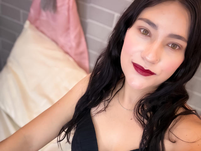 TamaraCians with long black hair, red lipstick, nose ring, and black dress, smiling at the camera, showing her small tits and fair skin. She's sitting on a bed.